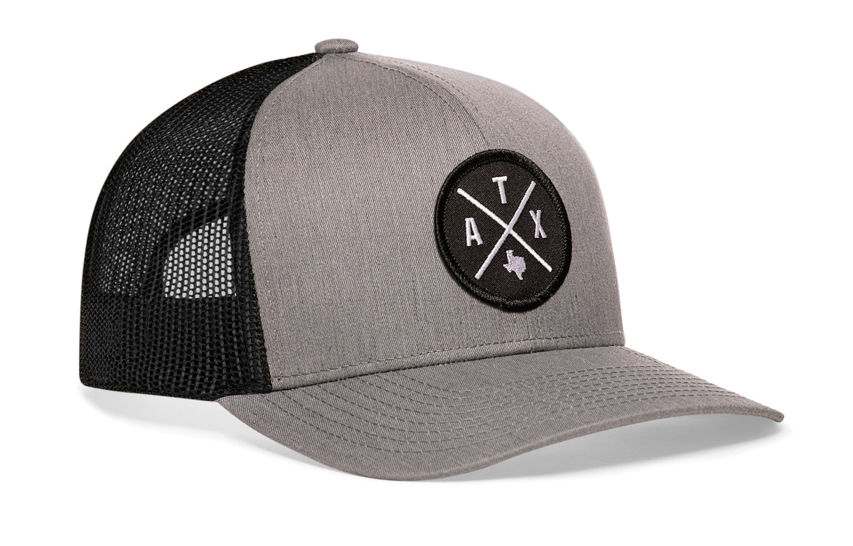 gray-black trucker hat viewed from the right side, showcasing a bold black and white embroidered X patch prominently displayed on the front
