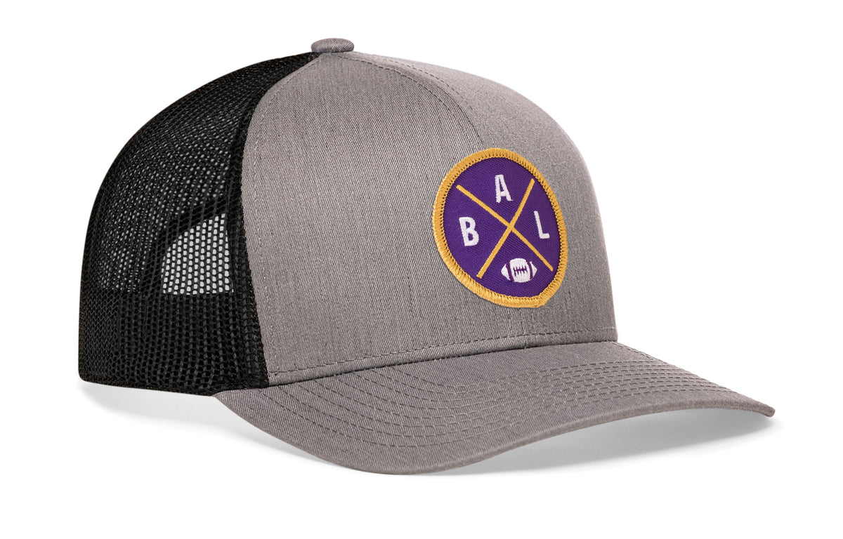gray-black trucker hat viewed from the right side, showcasing a bold purple and gold embroidered X patch prominently displayed on the front