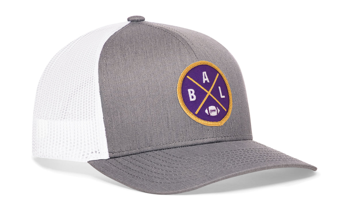 gray-white trucker hat viewed from the right side, showcasing a bold purple and gold embroidered X patch prominently displayed on the front