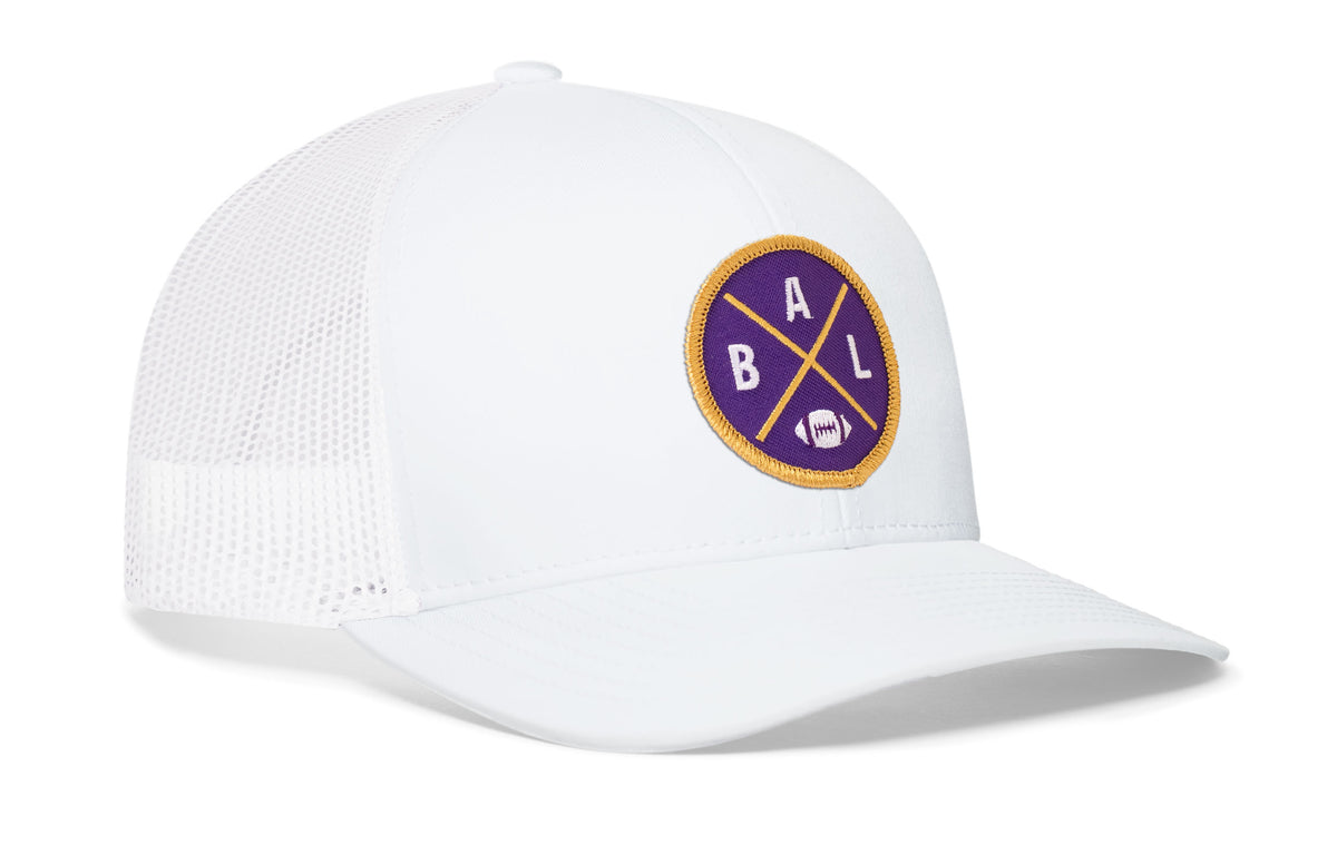 white trucker hat viewed from the right side, showcasing a bold purple and gold embroidered X patch prominently displayed on the front