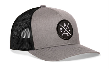 gray-black trucker hat viewed from the right side, showcasing a bold black and white embroidered X patch prominently displayed on the front