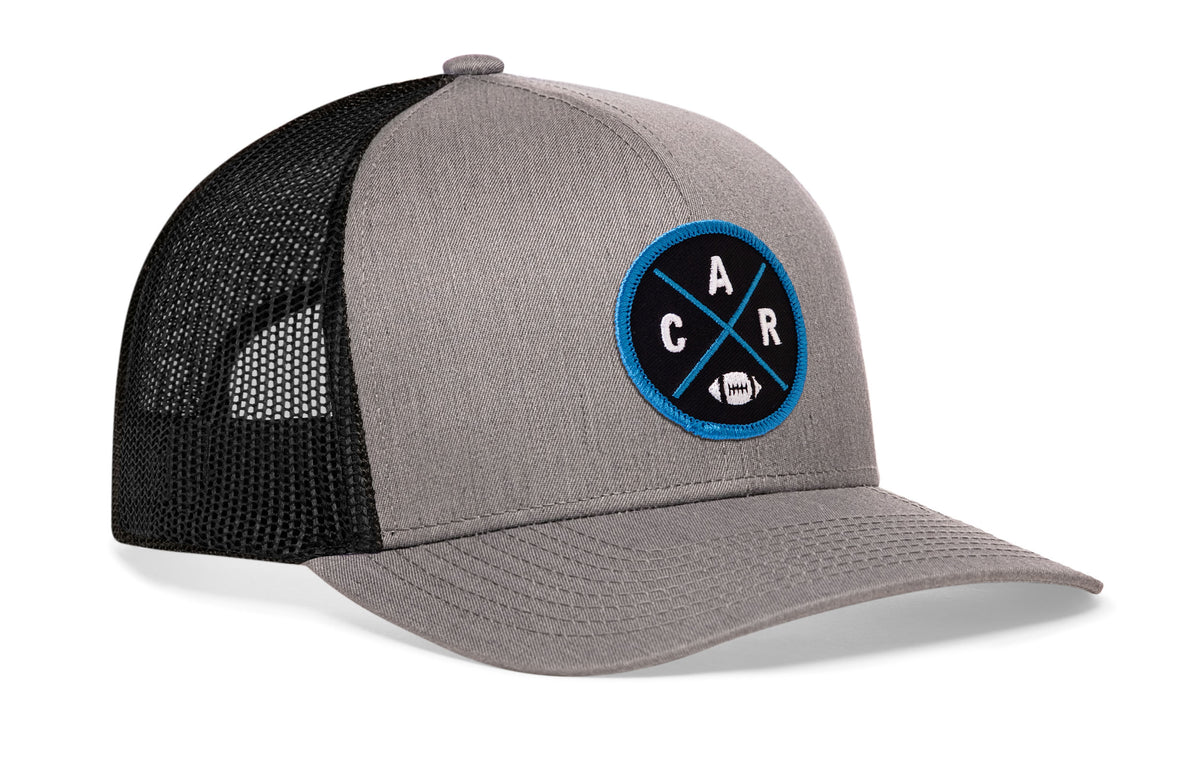 gray-black trucker hat viewed from the right side, showcasing a bold blue and black embroidered X patch prominently displayed on the front