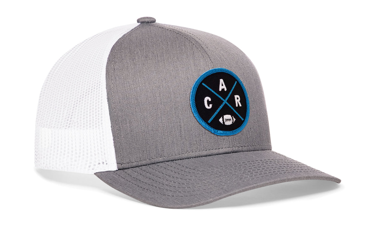 gray-white trucker hat viewed from the right side, showcasing a bold blue and black embroidered X patch prominently displayed on the front