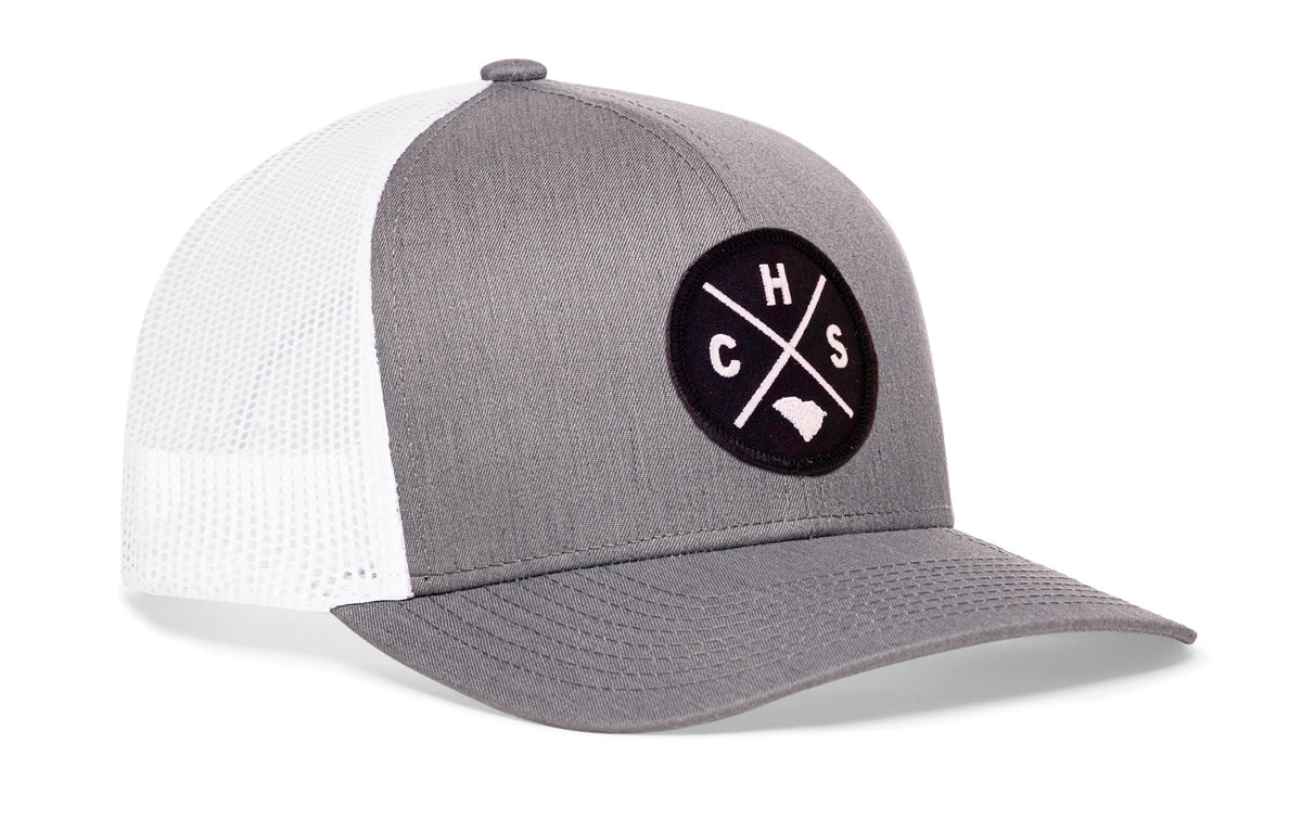 gray-white trucker hat viewed from the right side, showcasing a bold black and white embroidered X patch prominently displayed on the front