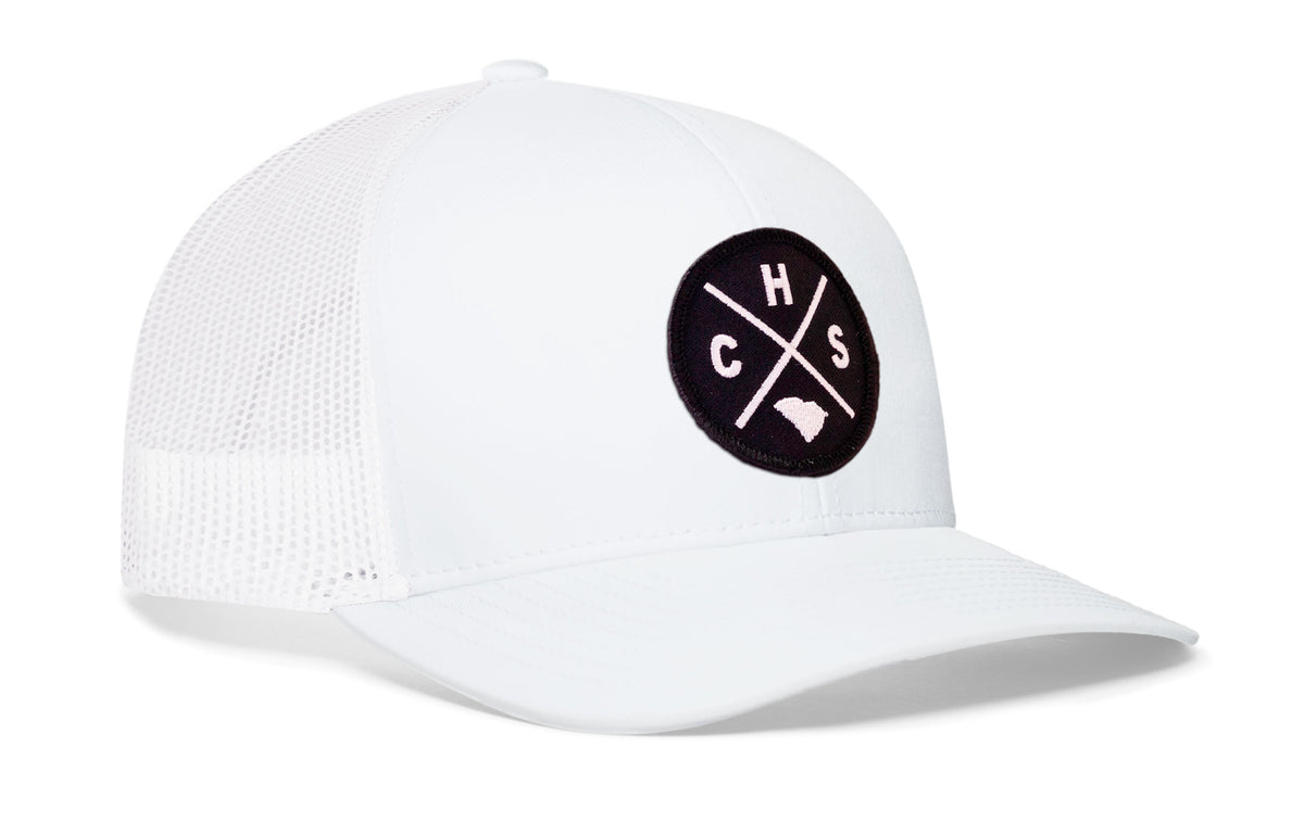 white trucker hat viewed from the right side, showcasing a bold black and white embroidered X patch prominently displayed on the front