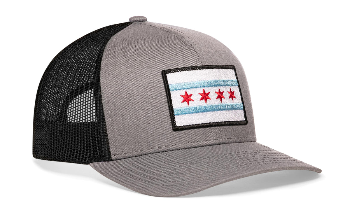 gray-black trucker hat viewed from the right side, showcasing a bold red and light blue embroidered flag patch prominently displayed on the front