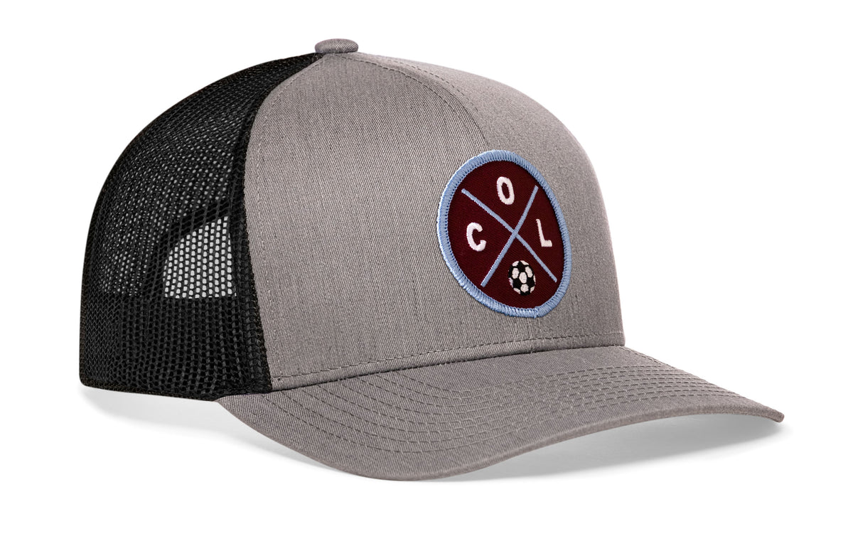 gray-black trucker hat viewed from the right side, showcasing a bold burgundy and blue embroidered X patch prominently displayed on the front
