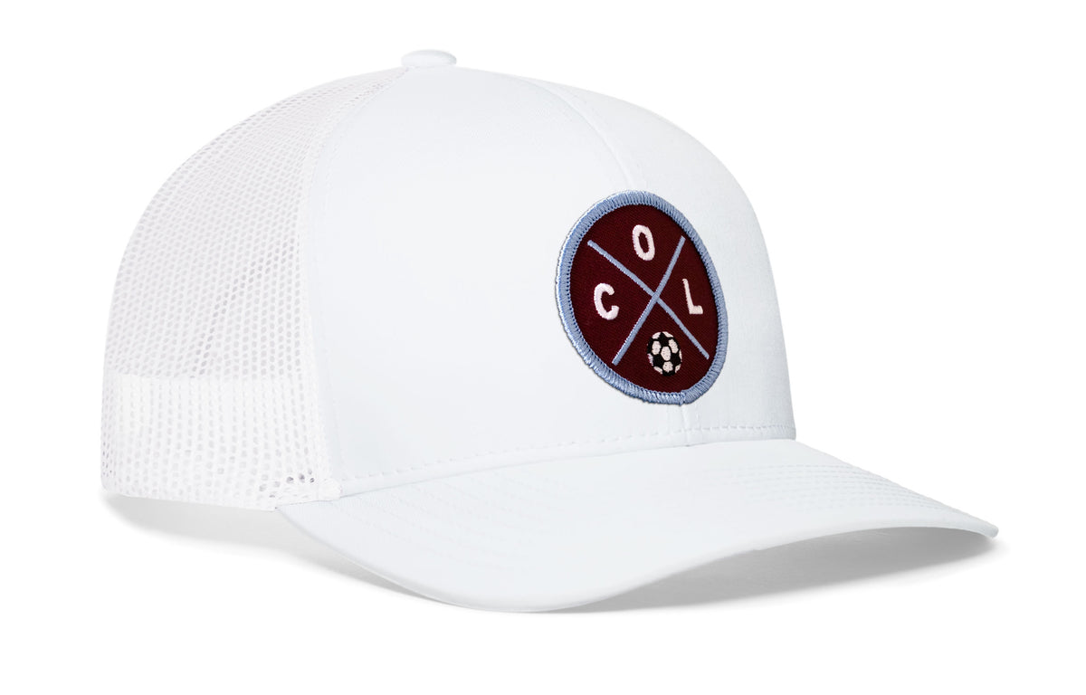white trucker hat viewed from the right side, showcasing a bold burgundy and blue embroidered X patch prominently displayed on the front