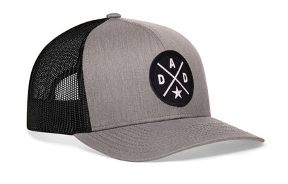 gray-black trucker hat viewed from the right side, showcasing a bold black and white embroidered X patch prominently displayed on the front