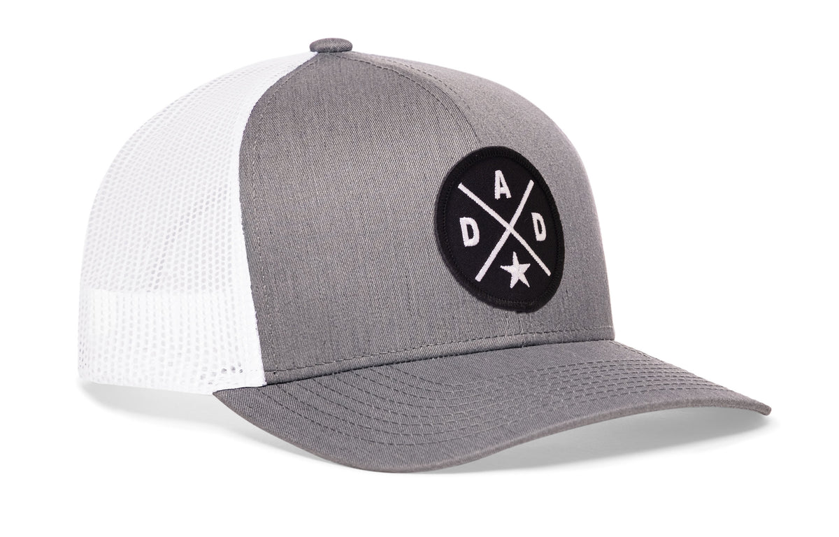 gray-white trucker hat viewed from the right side, showcasing a bold black and white embroidered X patch prominently displayed on the front