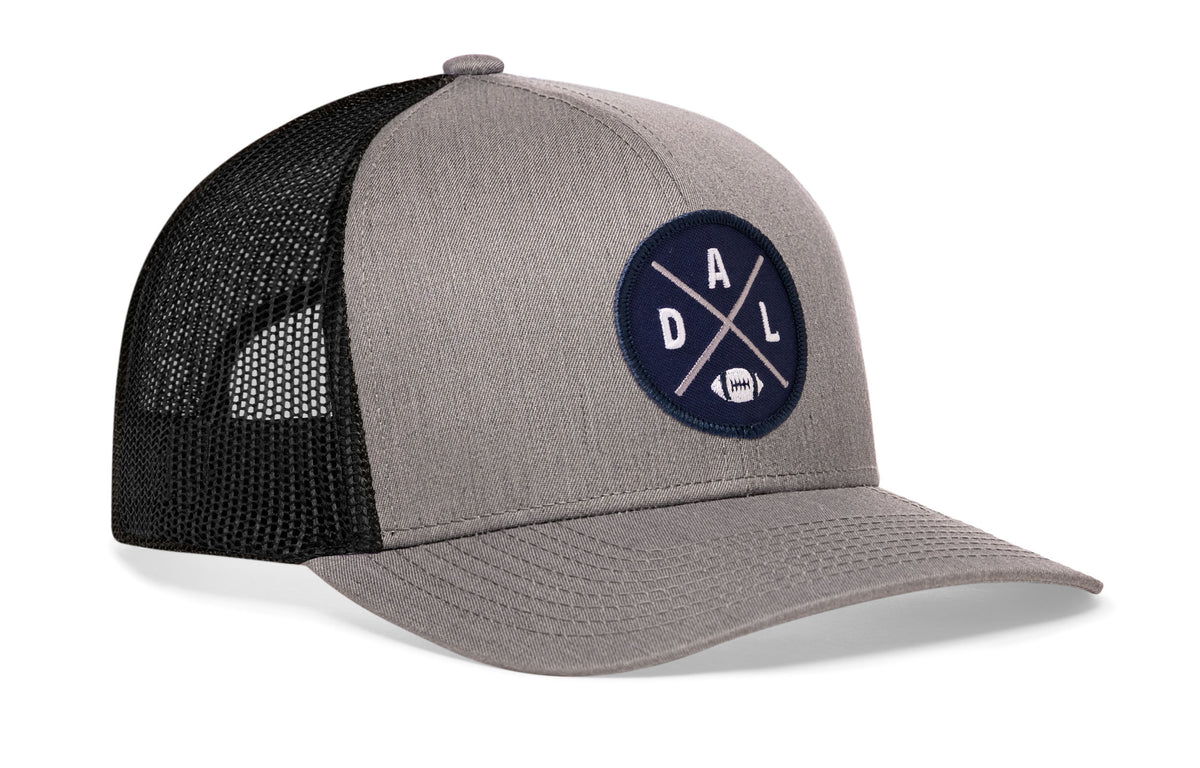 gray-black trucker hat viewed from the right side, showcasing a bold navy blue and silver embroidered X patch prominently displayed on the front