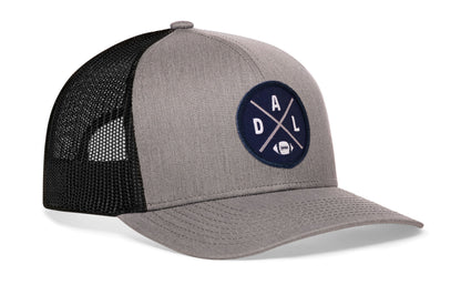 gray-black trucker hat viewed from the right side, showcasing a bold navy blue and silver embroidered X patch prominently displayed on the front