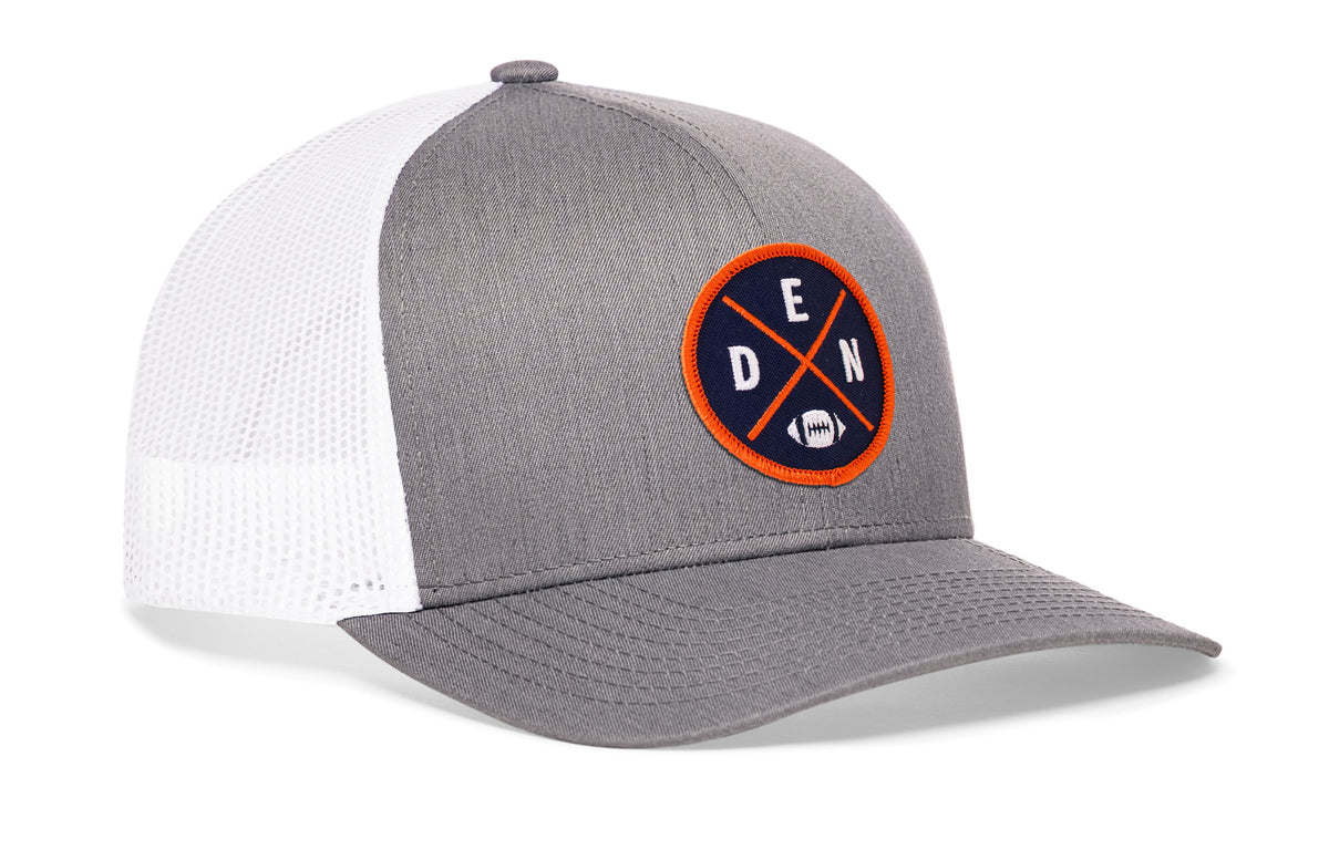 gray-white trucker hat viewed from the right side, showcasing a bold orange and navy embroidered X patch prominently displayed on the front