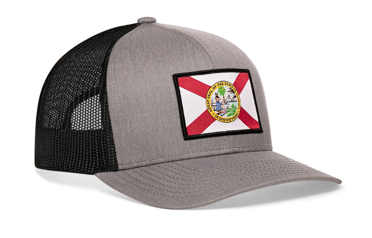gray-black trucker hat viewed from the right side, showcasing a bold red, white and yellow woven patch prominently displayed on the front