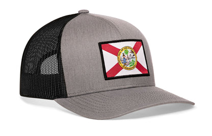 gray-black trucker hat viewed from the right side, showcasing a bold red, white and yellow woven patch prominently displayed on the front