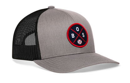 gray-black trucker hat viewed from the right side, showcasing a bold navy and red embroidered X patch prominently displayed on the front