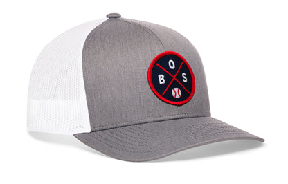 gray-white trucker hat viewed from the right side, showcasing a bold navy and red embroidered X patch prominently displayed on the front