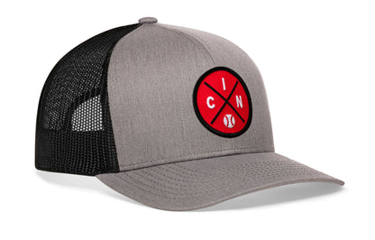 gray-black trucker hat viewed from the right side, showcasing a bold red and black embroidered X patch prominently displayed on the front