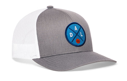 gray-white trucker hat viewed from the right side, showcasing a bold navy and royal embroidered X patch prominently displayed on the front