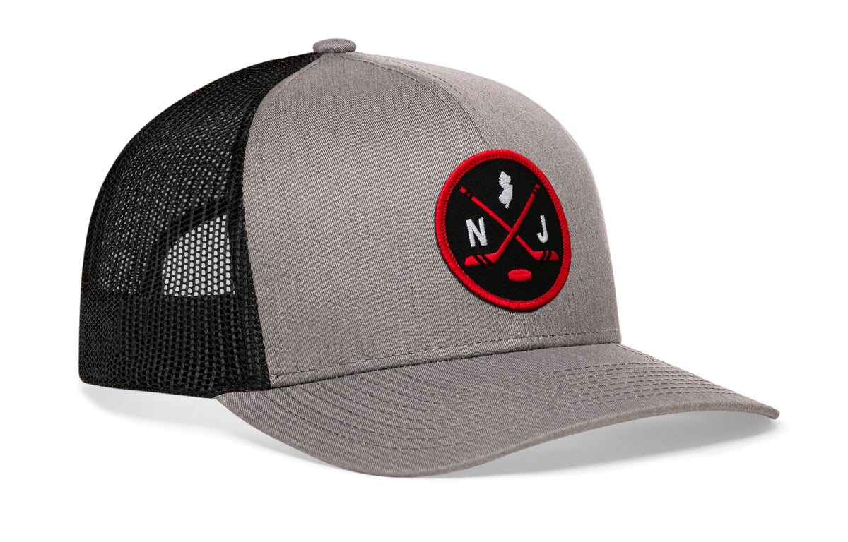 gray-black trucker hat viewed from the right side, showcasing a bold black and red embroidered X patch prominently displayed on the front