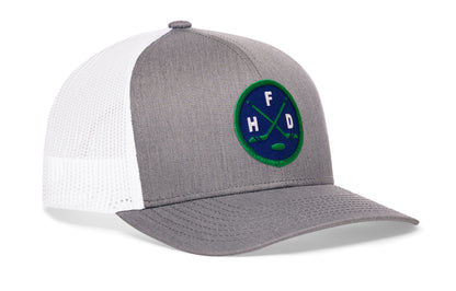 gray-white trucker hat viewed from the right side, showcasing a bold navy and green embroidered X patch prominently displayed on the front