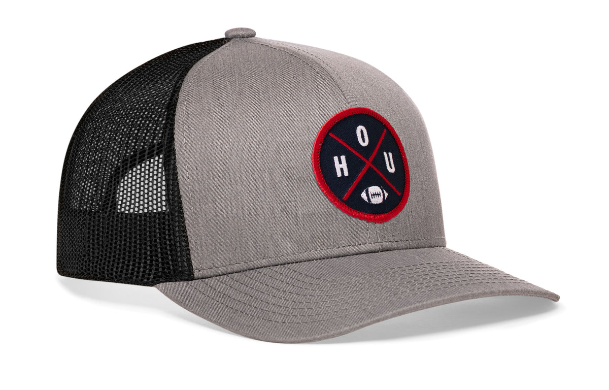gray-black trucker hat viewed from the right side, showcasing a bold navy blue and red embroidered X patch prominently displayed on the front
