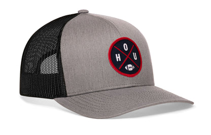 gray-black trucker hat viewed from the right side, showcasing a bold navy blue and red embroidered X patch prominently displayed on the front