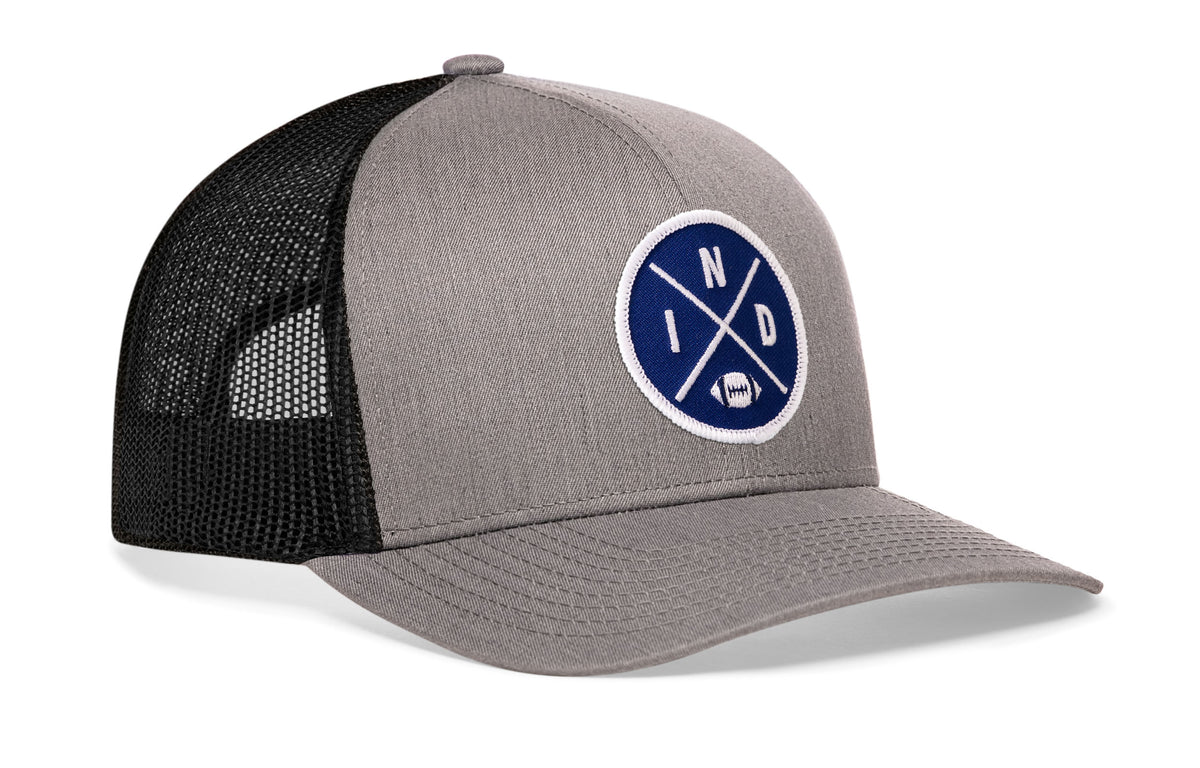 gray-black trucker hat viewed from the right side, showcasing a bold blue and white embroidered X patch prominently displayed on the front