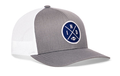 gray-white trucker hat viewed from the right side, showcasing a bold blue and white embroidered X patch prominently displayed on the front