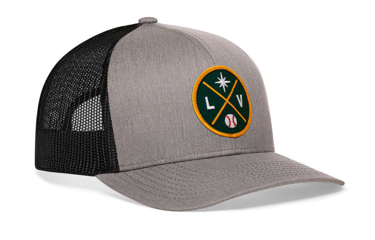 gray-black trucker hat viewed from the right side, showcasing a bold green and gold embroidered X patch prominently displayed on the front