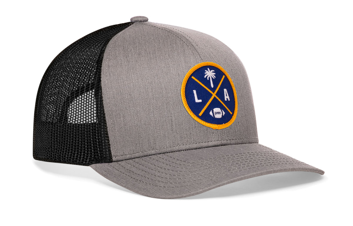 gray-black trucker hat viewed from the right side, showcasing a bold dark blue and yellow embroidered X patch prominently displayed on the front
