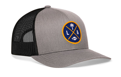 gray-black trucker hat viewed from the right side, showcasing a bold dark blue and yellow embroidered X patch prominently displayed on the front
