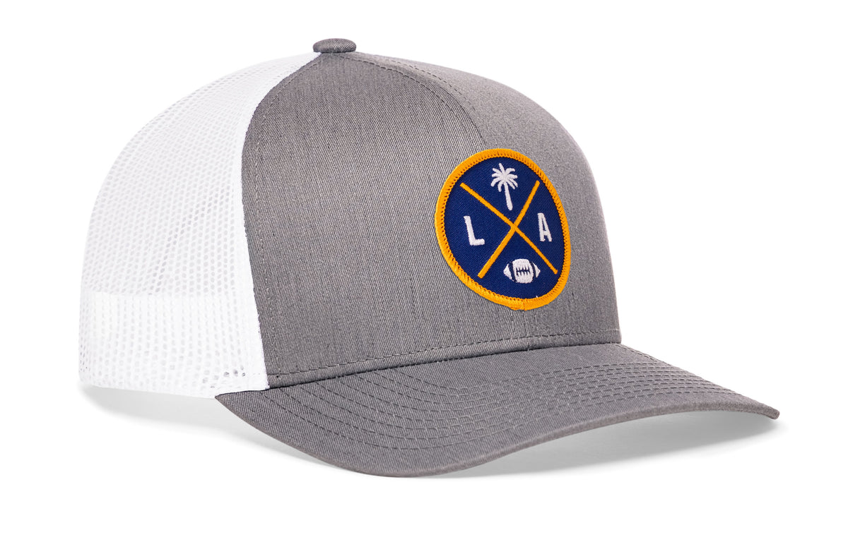 gray-white trucker hat viewed from the right side, showcasing a bold dark blue and yellow embroidered X patch prominently displayed on the front