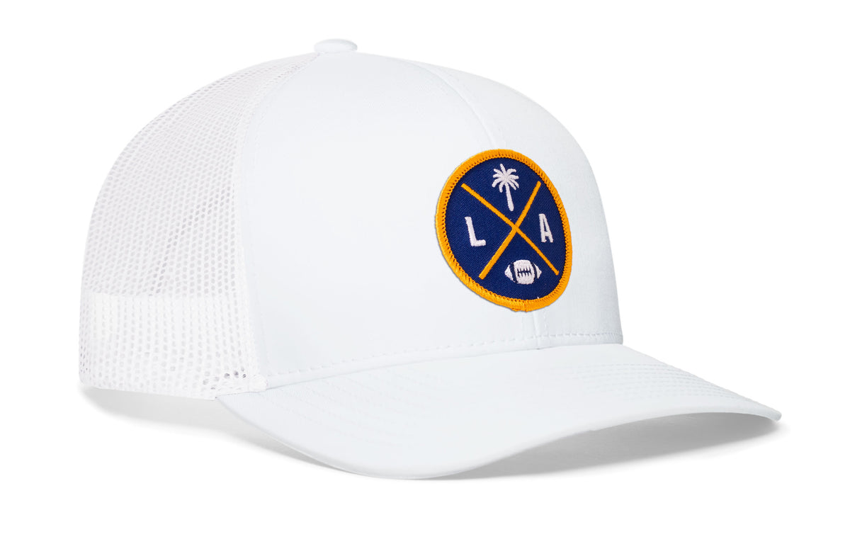white trucker hat viewed from the right side, showcasing a bold dark blue and yellow embroidered X patch prominently displayed on the front