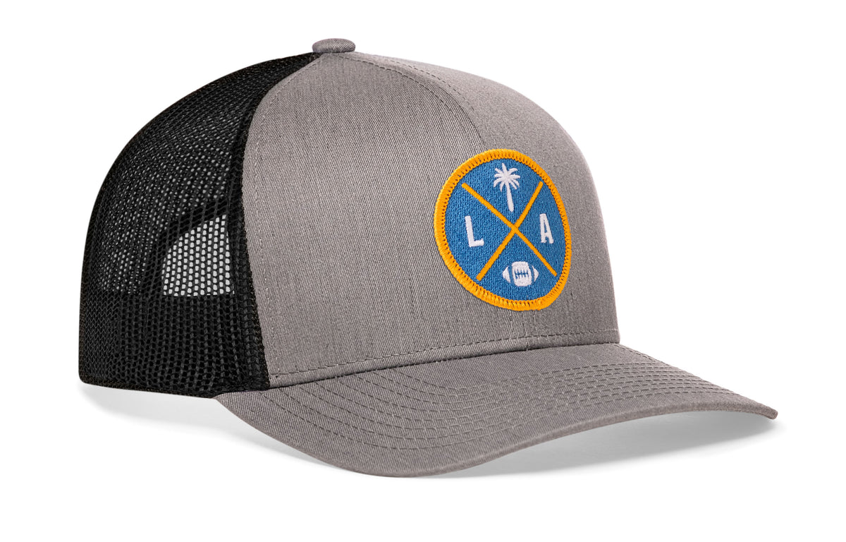 gray-black trucker hat viewed from the right side, showcasing a bold light blue and yellow embroidered X patch prominently displayed on the front
