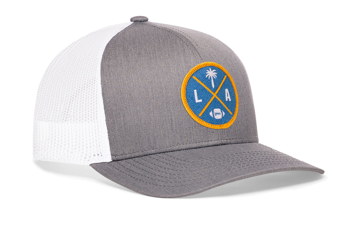 gray-white trucker hat viewed from the right side, showcasing a bold light blue and yellow embroidered X patch prominently displayed on the front