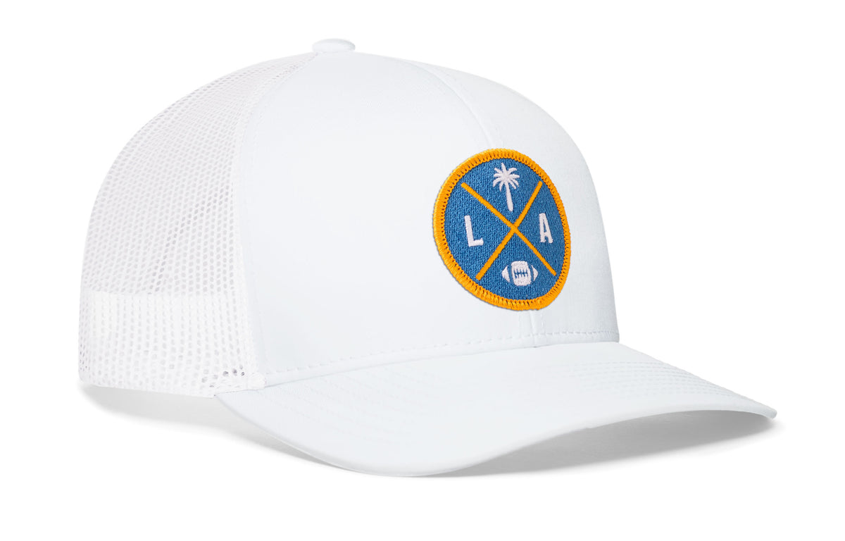 white trucker hat viewed from the right side, showcasing a bold light blue and yellow embroidered X patch prominently displayed on the front