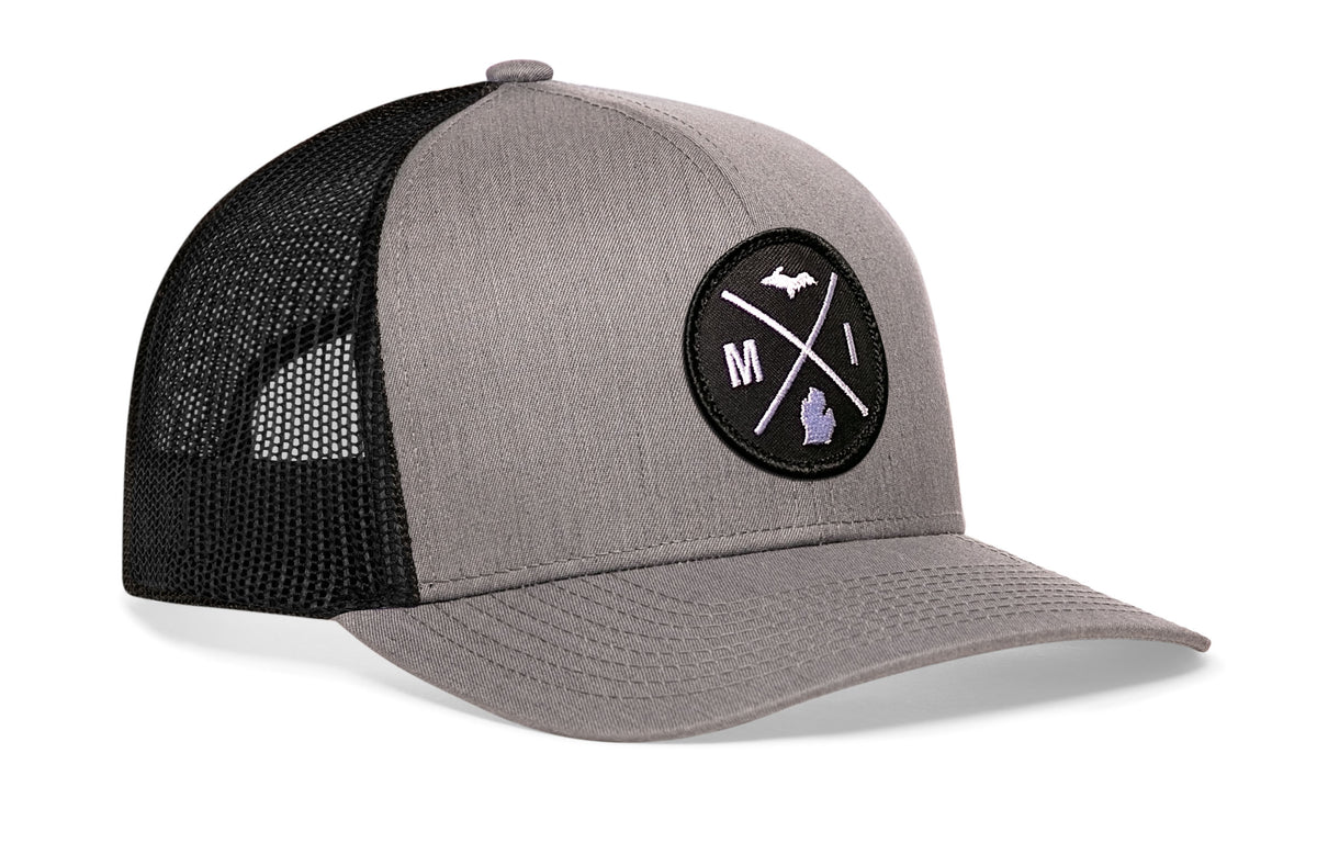 gray-black trucker hat viewed from the right side, showcasing a bold black and white embroidered X patch prominently displayed on the front