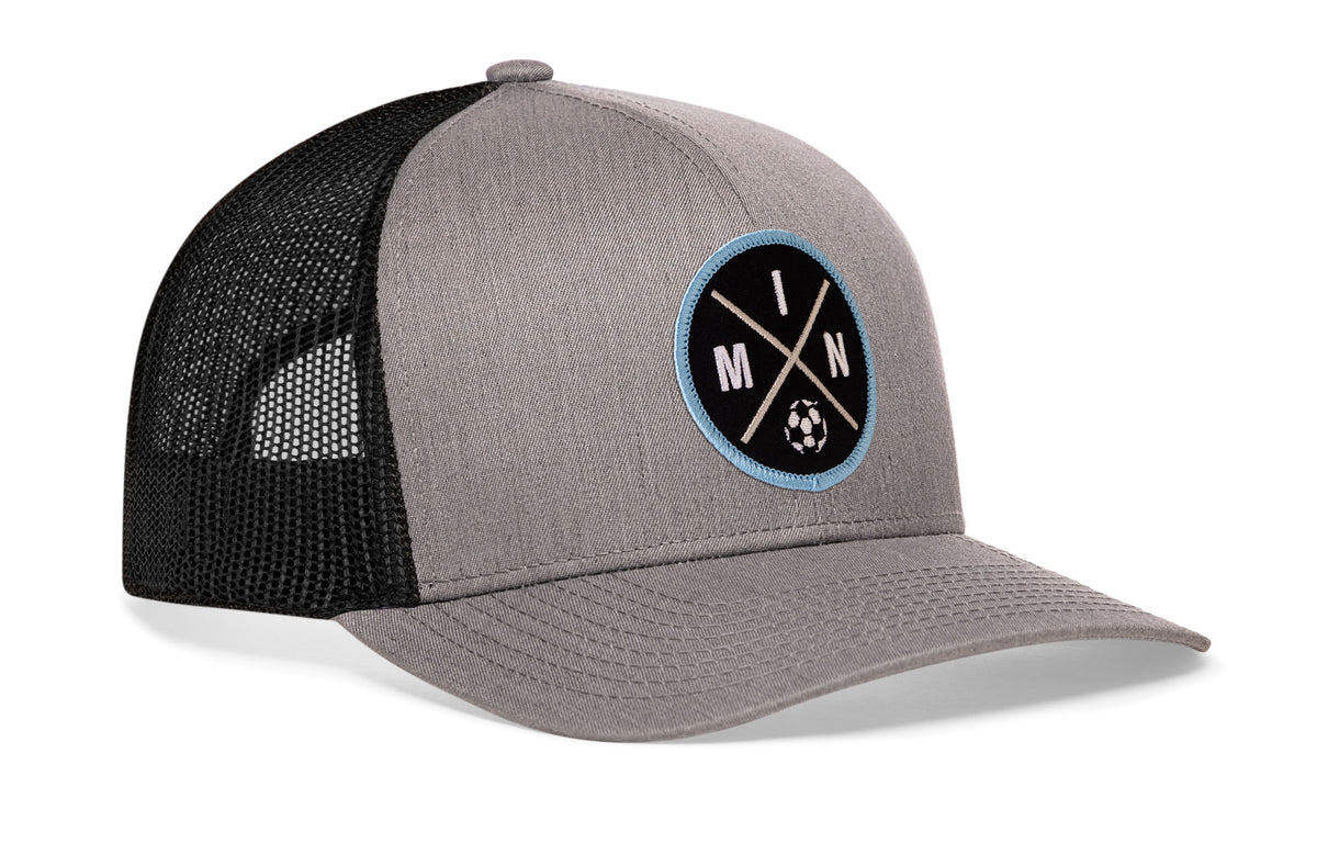 gray-black trucker hat viewed from the right side, showcasing a bold black and blue embroidered X patch prominently displayed on the front