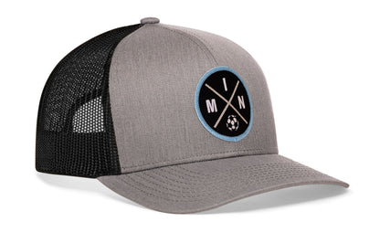 gray-black trucker hat viewed from the right side, showcasing a bold black and blue embroidered X patch prominently displayed on the front