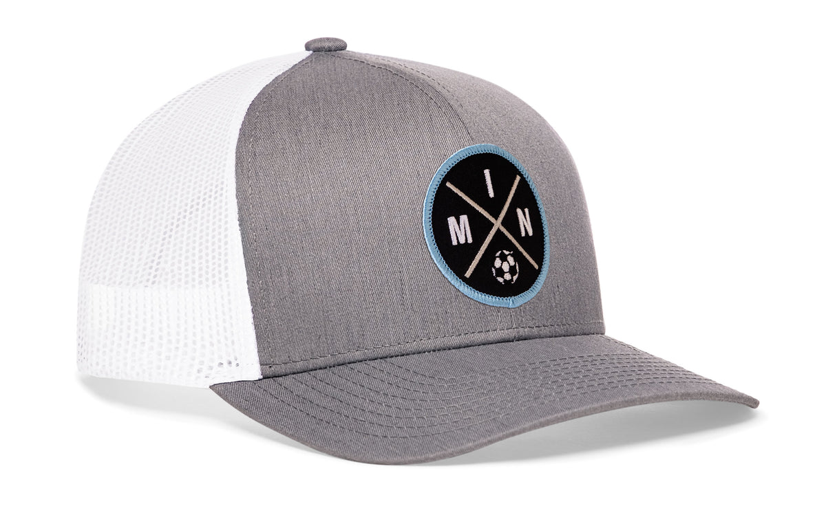gray-white trucker hat viewed from the right side, showcasing a bold black and blue embroidered X patch prominently displayed on the front