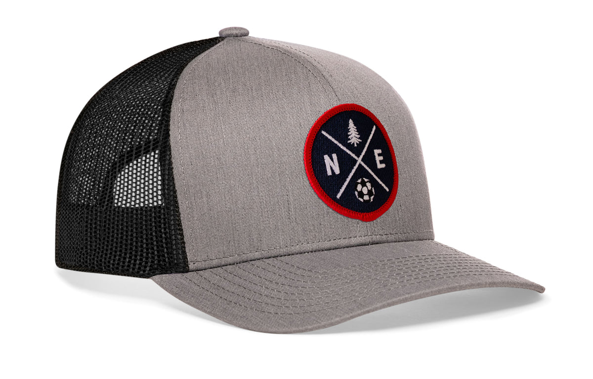 gray-black trucker hat viewed from the right side, showcasing a bold red and blue embroidered X patch prominently displayed on the front