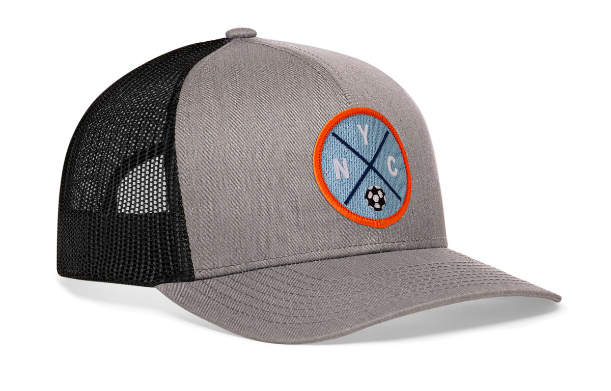 gray-black trucker hat viewed from the right side, showcasing a bold sky blue and orange embroidered X patch prominently displayed on the front