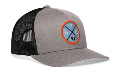 gray-black trucker hat viewed from the right side, showcasing a bold sky blue and orange embroidered X patch prominently displayed on the front