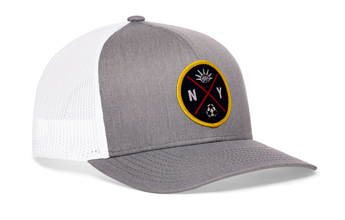 gray-white trucker hat viewed from the right side, showcasing a bold sky blue and orange embroidered X patch prominently displayed on the front