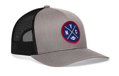 gray-black trucker hat viewed from the right side, showcasing a bold blue and red embroidered X patch prominently displayed on the front