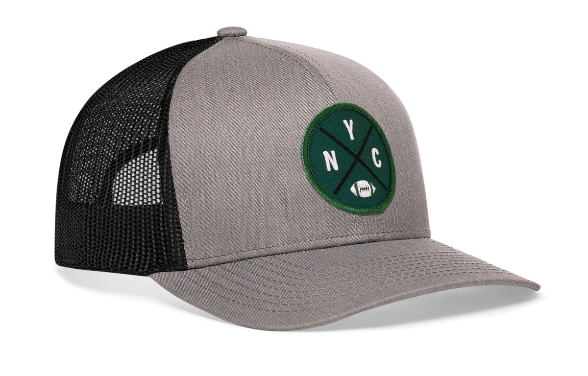 gray-black trucker hat viewed from the right side, showcasing a bold green, white and black embroidered X patch prominently displayed on the front