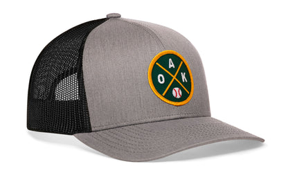 gray-black trucker hat viewed from the right side, showcasing a bold green and gold embroidered X patch prominently displayed on the front