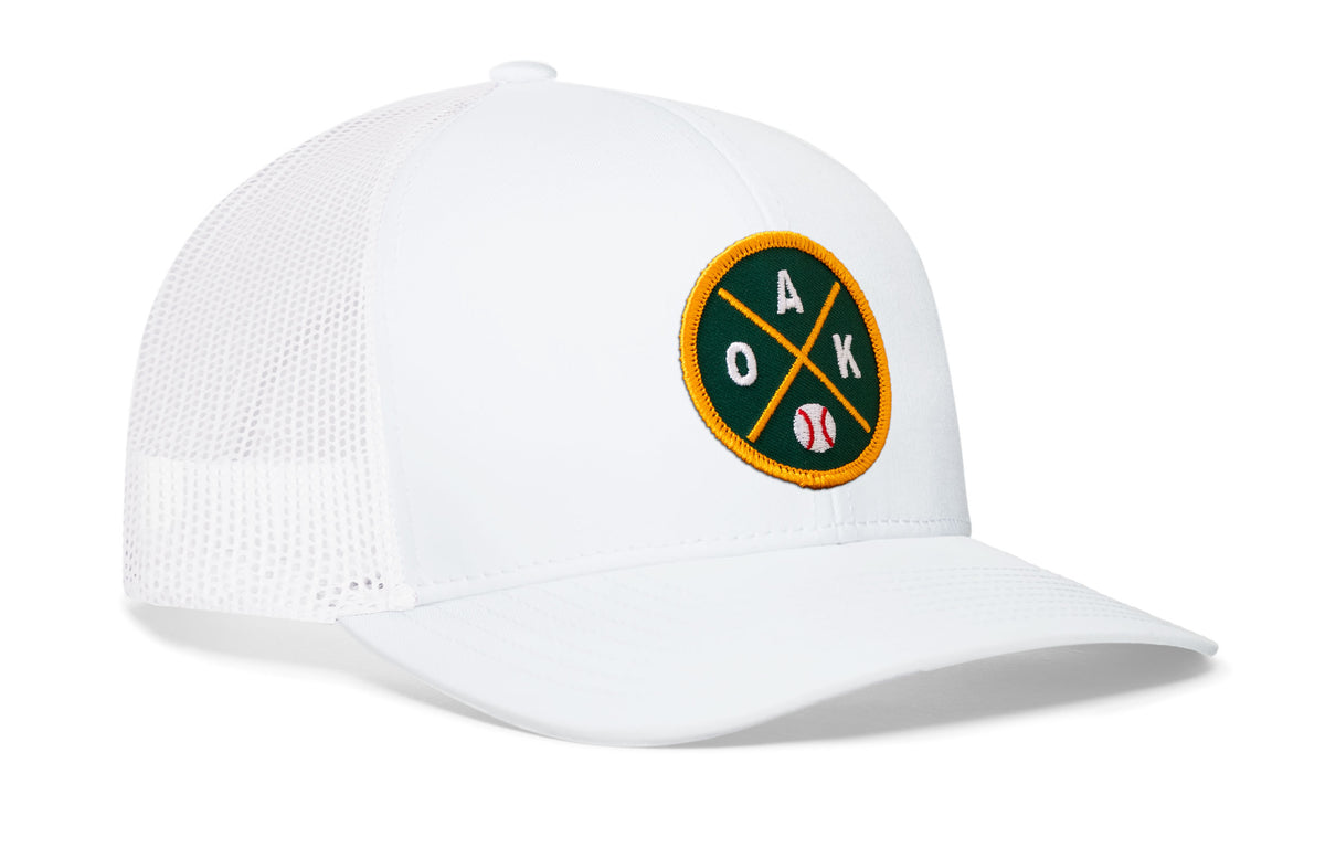 white trucker hat viewed from the right side, showcasing a bold green and gold embroidered X patch prominently displayed on the front