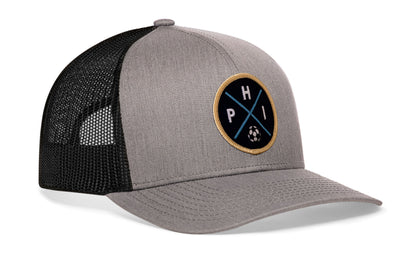 gray-black trucker hat viewed from the right side, showcasing a bold navy and gold embroidered X patch prominently displayed on the front